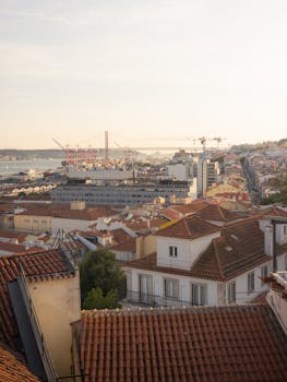 A beautiful panoramic view of Lisbon, Portugal featuring rooftops and the iconic 25 de Abril Bridge at sunset.
