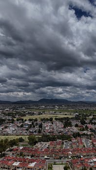 A dramatic aerial view of a Mexican city under a cloudy sky, showcasing urban landscape and distant mountains.