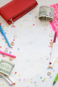 Flat lay of colorful stationery, wallet, and rolled dollar bills on marble background with copy space.