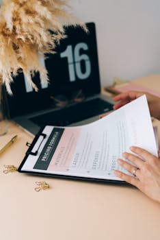 Hands review business documents on clipboard beside a laptop on a desk.